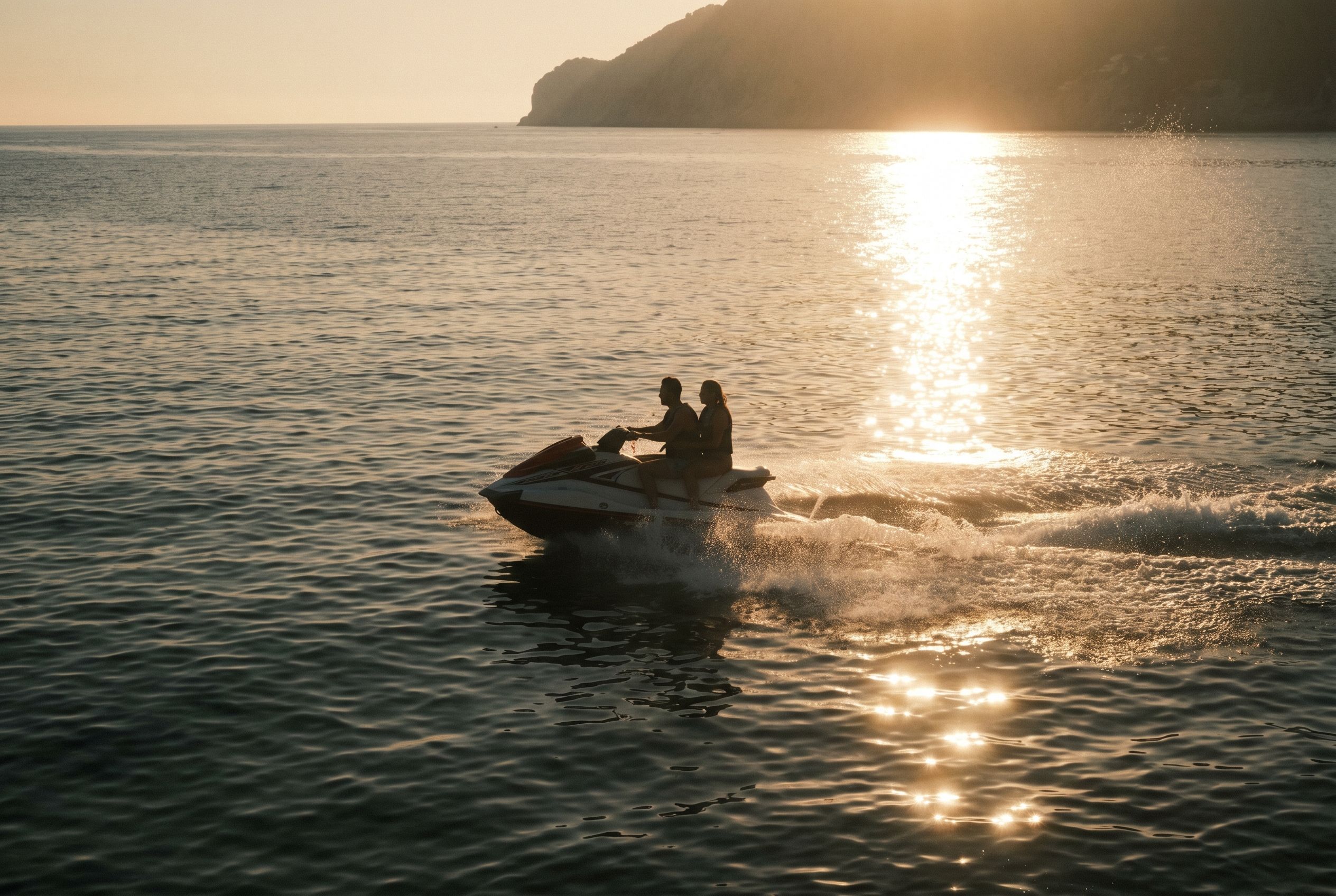 Two riders on a jet ski crossing a calm Mallorcan cove at golden hour